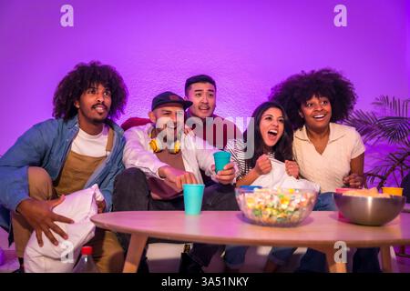 Un groupe diversifié de jeunes adultes est assis sur un canapé regardant un match de football avec pop-corn sous un éclairage LED violet. La scène de soirée de jeu en intérieur capture l'excitation, la célébration et le divertissement social. Banque D'Images