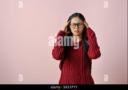 Une étudiante asiatique stressée massant ses tempes, suggérant un mal de tête ou une migraine. Un moment pensif sur le campus, captant la pression des études et la vie universitaire. Banque D'Images