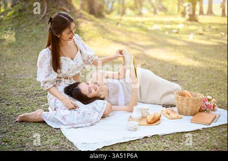 Femme asiatique souriante assise sur une couverture dans un parc ensoleillé, reposant un livre sur ses genoux avec sa petite amie à côté d'elle. Ils profitent d'un pique-nique de jour détendu en plein air, mettant en valeur la convivialité et les loisirs. Idéal pour le couple, le style de vie et les thèmes d'activités de plein air. Banque D'Images