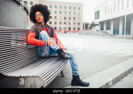 Un bel étudiant égyptien aux cheveux bouclés est assis sur un banc dans un cadre urbain, portant un pull orange vif et un Jean. Le moment capture un style extérieur décontracté et des pauses d'étude entre les leçons. Banque D'Images