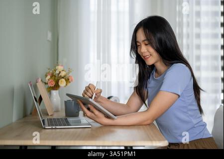 Belle jeune femme asiatique utilisant une tablette numérique à une table à l'intérieur. L'image met en évidence la technologie, l'apprentissage en ligne et la connectivité dans un environnement domestique. Convient au travail à distance, aux études et aux modes de vie contemporains. Banque D'Images