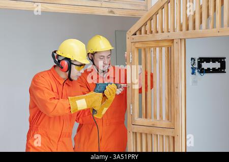 Les travailleurs sérieux dans les combinaisons de construction orange et les casques de sécurité installent des composants de construction en bois à l'intérieur d'une maison. Le travail d'équipe ciblé et l'équipement de protection reflètent les pratiques professionnelles des chantiers de construction. Idéal pour les projets, la menuiserie et les thèmes de reconstruction. Banque D'Images