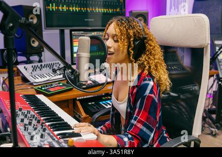 Une femme hispanique est assise dans un studio d'enregistrement jouant un synthétiseur et chantant dans un microphone. La configuration présente la production musicale et la performance vocale avec un équipement studio moderne. Banque D'Images