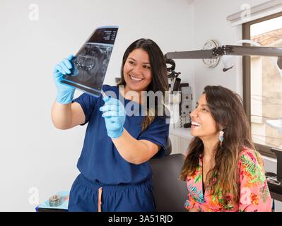 Une dentiste métisse souriante montre une radiographie dentaire panoramique à une patiente hispanique dans une clinique. Idéal pour les soins dentaires, la dentisterie et les communications de santé impliquant le travail d'équipe et l'orientation des patients. Banque D'Images