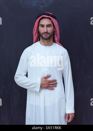 Portrait d'un jeune homme du moyen-Orient vêtu de vêtements traditionnels devant un tableau noir. L'image mélange la mode islamique moderne avec les concepts de Ramadan Kareem, adaptés aux campagnes culturelles ou religieuses. Un cadre studio et une expression confiante mettent en valeur le style contemporain dans la tradition. Banque D'Images