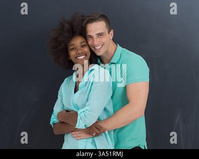 Femme noire souriante avec un Afro est embrassée par derrière par son petit ami caucasien dans un studio à côté d'un tableau noir. Ce concept de couple diversifié et multiculturel est idéal pour le style de vie, les relations et les visuels de travail d'équipe. Banque D'Images