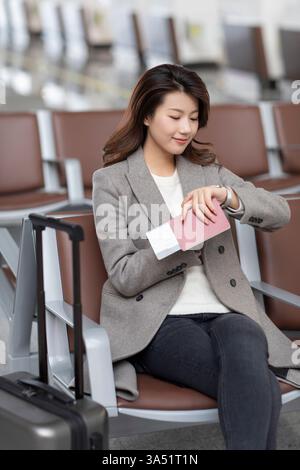 Femme chinoise regardant sa montre tenant passeport et billet attendant le vol assis avec les jambes croisées sur le banc à l'aéroport Banque D'Images