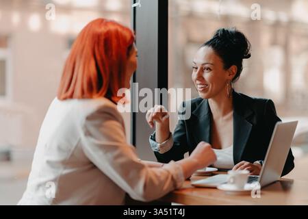 Femme d'affaires hispanique souriante en costume noir discutant des plans d'affaires avec une collègue féminine caucasienne tout en étant assise à table avec un café et un lapt... Banque D'Images