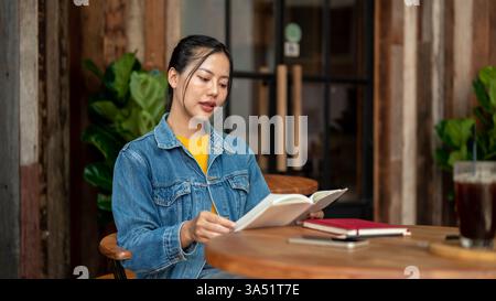 Femme asiatique dans une veste en denim assise à une table dans un café, absorbée dans un livre. Cette scène convient aux moments d'étude ou d'apprentissage dans un café décontracté ou un espace de coworking. Banque D'Images