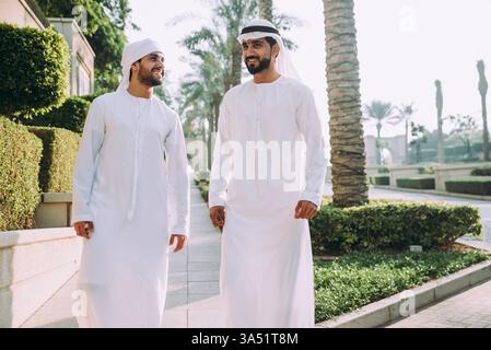 Deux jeunes hommes d'affaires en tenue de kandura marchant le long de Dubai Marina. La scène urbaine capture l'amitié et le style professionnel au moyen-Orient. Idéal pour les voyages d'affaires, le réseautage et le contenu régional de style de vie d'entreprise. Banque D'Images