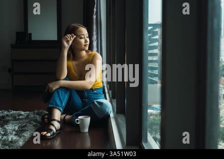 Jeune femme asiatique debout près d'une fenêtre avec une tasse, profitant d'une matinée tranquille à la maison. L'intérieur détendu et confortable évoque le confort, la vie urbaine et le temps d'arrêt paisible. Idéal pour le contenu sur les rituels du café, l'auto-soin et la vie en ville. Banque D'Images