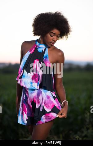 Portrait d'une femme afro-américaine souriante dans une robe d'été par une chaude soirée. Une image de style de vie élégante capturant le maquillage naturel et les cheveux bouclés pour la mode et les traits de beauté. Idéal pour la mode, le style de vie et les campagnes marketing. Banque D'Images
