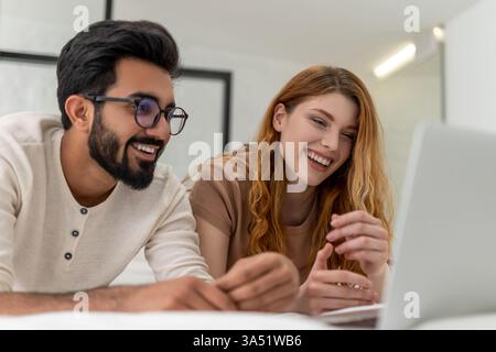 Mari indien souriant portant des lunettes regardant un film sur ordinateur portable avec une femme caucasienne couchée sur le lit à la maison Banque D'Images