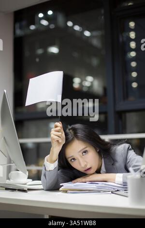 Femme d'affaires chinoise travaillant tard dans un bureau moderne. La scène met en évidence les heures supplémentaires et la concentration professionnelle avec un ordinateur et la paperasse sur le bureau. Convient pour les articles sur la vie d'entreprise, le stress au travail et la productivité des heures supplémentaires. Banque D'Images
