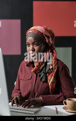 Femme musulmane africaine en hijab dans un bureau regardant un écran d'ordinateur et dactylographiant. Un portrait moderne et professionnel mettant en valeur la diversité et la technologie en milieu de travail. Idéal pour les entreprises, la diversité et les campagnes inclusives sur le lieu de travail. Banque D'Images