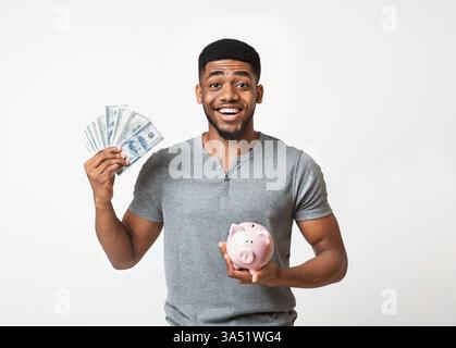 Heureux homme afro-américain tenant une pile d'argent et une tirelire sur un fond blanc. Cette image sur le thème de la finance est idéale pour les campagnes d'épargne, de budgétisation et d'investissement. Idéal pour les opérations bancaires, la planification financière et le contenu patrimonial avec un sentiment positif et optimiste. Banque D'Images