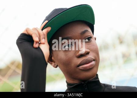 Portrait d'une femme afro-américaine élégante portant une casquette, regardant loin avec une ambiance décontractée et confiante. Une photo de style de vie urbain adaptée aux campagnes de mode et streetwear, avec des allusions de ville en toile de fond. Banque D'Images