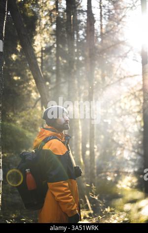 Vue de côté d'un voyageur barbu en vêtements chauds avec un sac à dos, debout dans une forêt d'automne dans le parc national de Stockholm, Suède. Les paysages ensoleillés suggèrent l'exploration et l'errance, idéales pour les voyages et les thèmes d'aventure en plein air. Banque D'Images