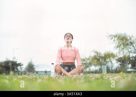 Femme athlétique calme en vêtements d'activité est assis sur le sol avec les yeux fermés, pratiquant Padmasana à côté d'une bouteille d'eau. Cette image de yoga et de méditation communique bien-être, pleine conscience et équilibre. Idéal pour les campagnes de bien-être, de fitness et de mode de vie sain. Banque D'Images