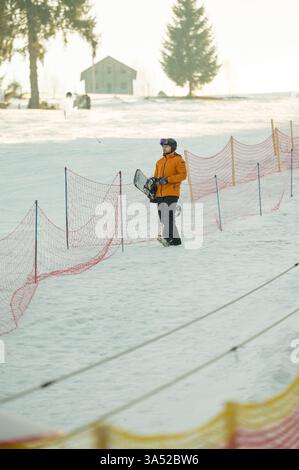 Avec un équipement orange éclatant, un snowboarder se tient au bord d'une pente enneigée, prêt à affronter la course à venir dans un paysage hivernal tranquille. Banque D'Images