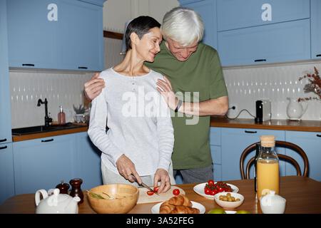 Couple senior préparant avec amour le petit déjeuner ensemble dans une cuisine moderne avec des armoires bleues et divers ingrédients frais sur le comptoir Banque D'Images
