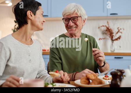 Deux aînés partageant le petit déjeuner dans un cadre de cuisine confortable, souriant et à l'air détendu. Les deux sont engagés dans une conversation heureuse tout en profitant de leur repas Banque D'Images