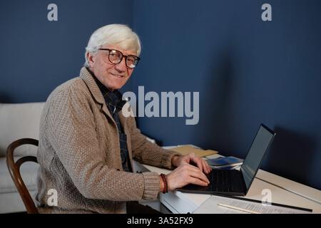 Homme âgé avec des lunettes à l'aide d'un ordinateur portable au bureau à la maison, assis sur une chaise en bois avec une pile de livres à proximité, et souriant chaleureusement Banque D'Images