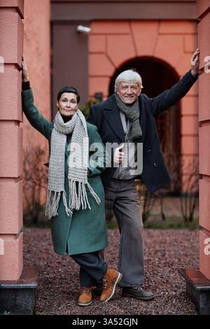 Couple de personnes âgées souriantes debout à l'extérieur près de l'entrée du bâtiment, les deux portant une tenue d'hiver, affichant chaleur et bonheur dans un environnement serein Banque D'Images
