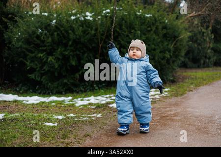 Un tout-petit joyeux, vêtu d'une combinaison de neige bleu vif et d'un chapeau chaud, explore joyeusement un chemin en plein air enneigé, brandissant un petit bâton pendant cette belle Banque D'Images
