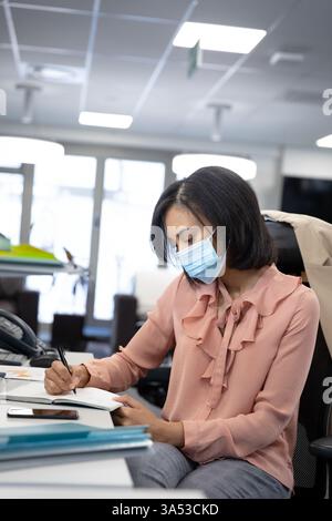 Femme professionnelle portant masque écrivant des notes au bureau, concentré sur le travail Banque D'Images