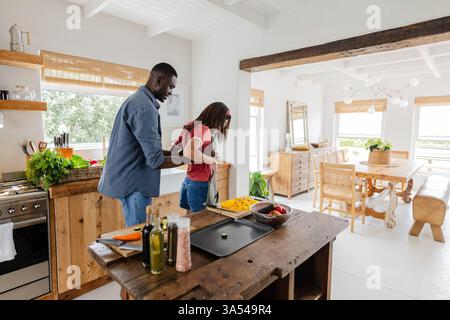 Couple diversifié cuisinant ensemble dans une cuisine lumineuse, préparant des légumes frais pour le déjeuner Banque D'Images