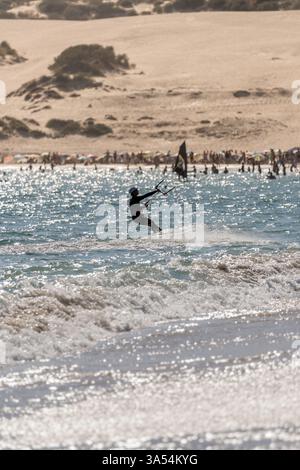 Silhouette d'un kite surfeur chevauchant les vagues au coucher du soleil, ralentissant pour atteindre le rivage d'une plage pleine de baigneurs et les dunes en arrière-plan en Banque D'Images