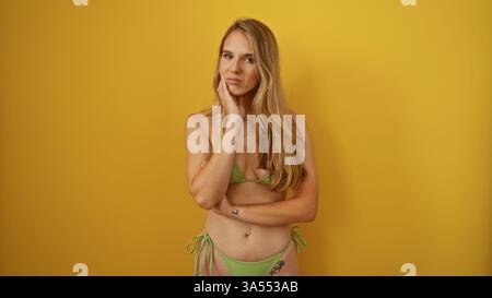 Belle jeune femme aux longs cheveux blonds dans un bikini vert posant contre un mur jaune vif, véhiculant une expression réfléchie et un ensemble isolé Banque D'Images