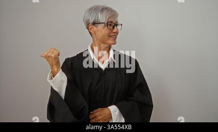 Femme aux cheveux courts gris portant des lunettes pointe avec son pouce sur le côté sur un fond blanc, exprimant confiance et maturité. Banque D'Images