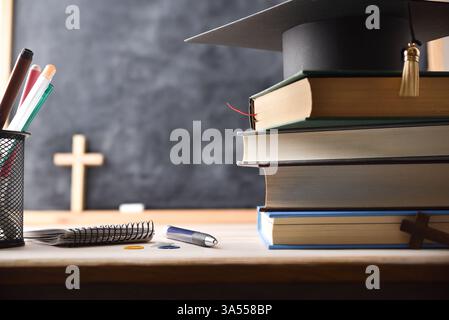 Pile de livres éducatifs et un mortarboard sur une table avec des outils et un fond avec un tableau noir et une croix en bois. Concept d'études religieuses. Avant Banque D'Images