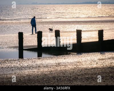 Silhouette d'une personne promenant son chien sur une plage vide avec la marée à Camber Sands Banque D'Images