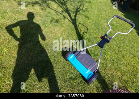 Ombre d'une personne debout près d'une tondeuse à gazon sur une pelouse verte pendant une journée ensoleillée, suggérant des soins de jardin et une activité de plein air. Banque D'Images