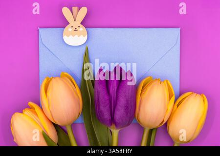 Nature morte de printemps avec une enveloppe bleue bébé, un clip en forme de lapin et des tulipes sur un fond rose vif, parfait pour les dessins de Pâques. Banque D'Images