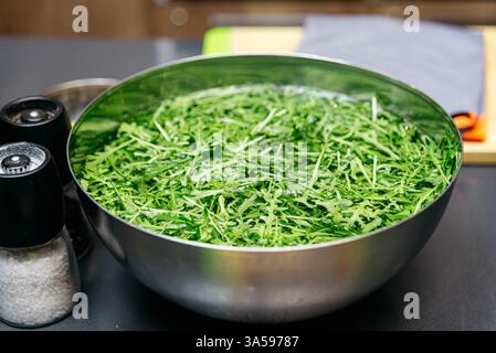 Un bol en acier inoxydable rempli de feuilles de roquette fraîches trempant dans l'eau, placé sur un comptoir de cuisine avec salière et poivrière à proximité. Banque D'Images