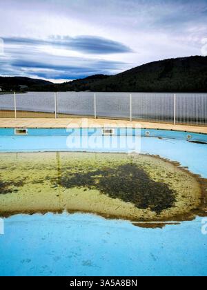 Une piscine négligée avec de l'eau trouble se trouve vide, encadrée par une clôture en fil de fer et un lac tranquille en arrière-plan, dans un ciel nuageux Banque D'Images