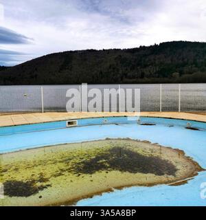 Une piscine négligée avec de l'eau trouble se trouve vide, encadrée par une clôture en fil de fer et un lac tranquille en arrière-plan, dans un ciel nuageux Banque D'Images