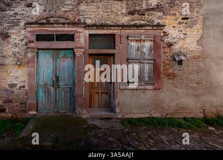 Double porte en bois rouge vieilli avec vitres brisées et grilles verticales en fer, dans un mur rouge altéré. Banque D'Images
