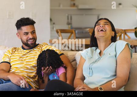 Famille heureuse se relaxant sur le canapé, mère riant tandis que sa fille couvre le visage de manière ludique Banque D'Images