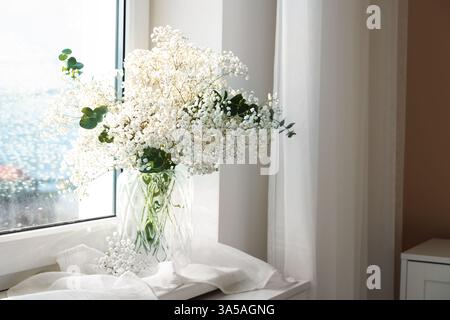 Bouquet de belles fleurs de gypsophila et branches d'eucalyptus dans un vase sur le rebord de la fenêtre à l'intérieur Banque D'Images