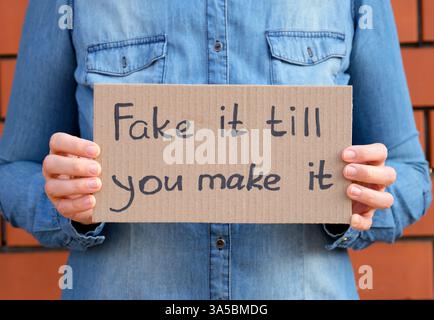Femme tenant un morceau de carton avec phrase Fake It jusqu'à ce que vous le fassiez contre un fond de mur de briques. Banque D'Images
