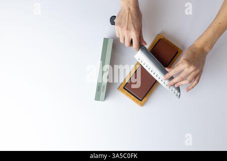 top shot sur fond blanc sur une table, les mains des femmes aiguisent un couteau en acier, une serrure sous le bras, une protection contre les coupures, une pierre rouge moyennement dure Banque D'Images
