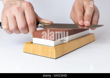 gros plan sur fond blanc sur une table, les mains de l'homme affûtent une protection de couteau en acier contre les coupures, une pierre rouge moyennement dure pour affûter les couteaux Banque D'Images