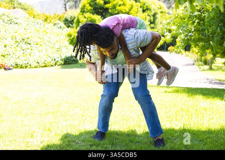 Père donnant à sa fille un tour de piggyback dans le parc ensoleillé, les deux riant ensemble Banque D'Images