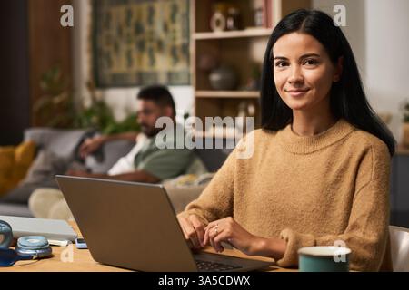 Portrait de femme avec de longs cheveux noirs à l'aide d'un ordinateur portable à la maison avec un homme en arrière-plan affichant une atmosphère confortable et détendue Banque D'Images