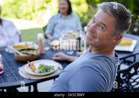 Homme souriant profitant d'un déjeuner en plein air avec des amis seniors, se relaxant dans le cadre du jardin Banque D'Images